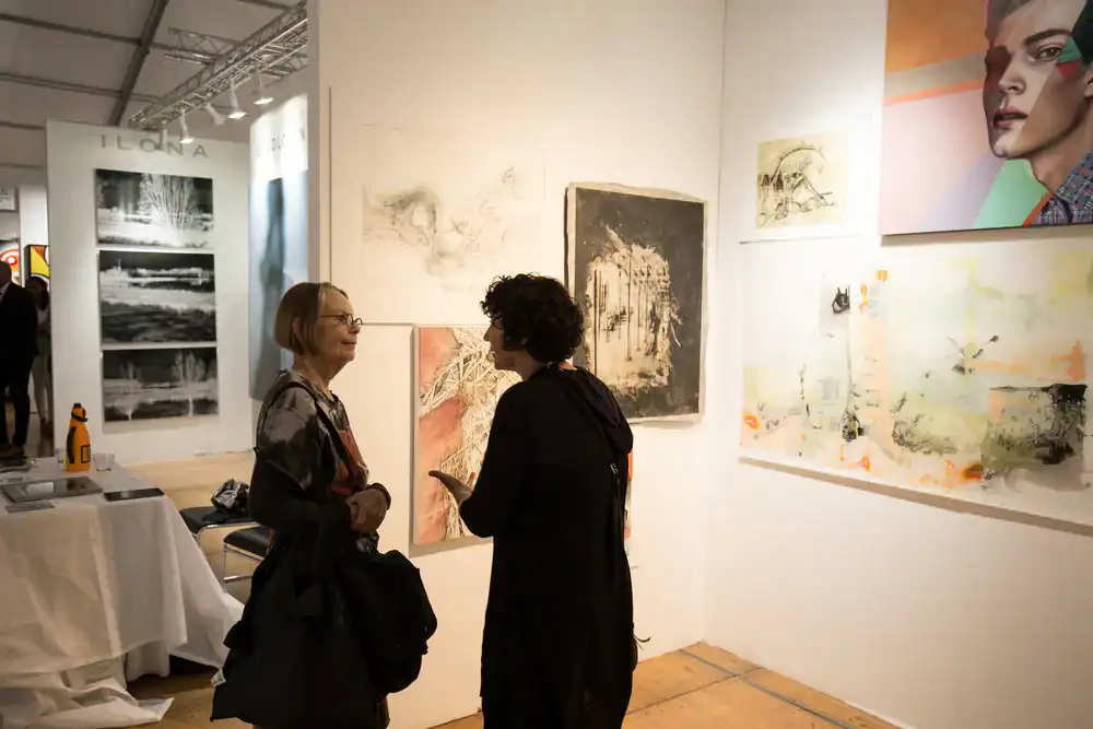 Two women stand talking in front of a white wall displaying various artworks at an art gallery or exhibition. One table with books and a chair is visible on the left. The scene is warmly lit.