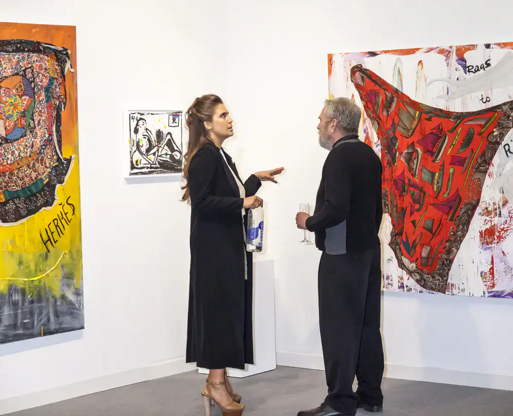 A woman and a man stand in an art gallery, engaged in conversation. Colorful abstract paintings hang on the white walls around them. The woman gestures with her hand, and the man holds a drink.