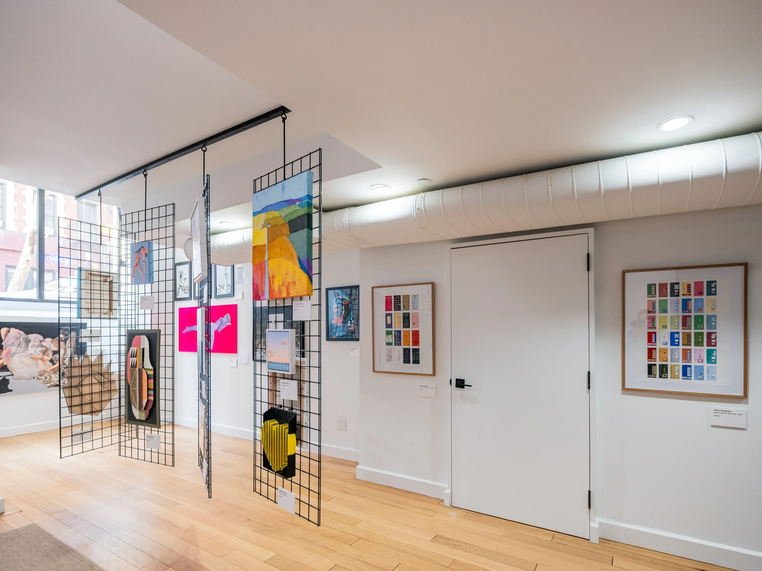 A modern art gallery room with wooden floors, white walls, and a white door. Metal grid panels hang from the ceiling, displaying colorful paintings and artworks. Framed art pieces are also mounted on the walls.