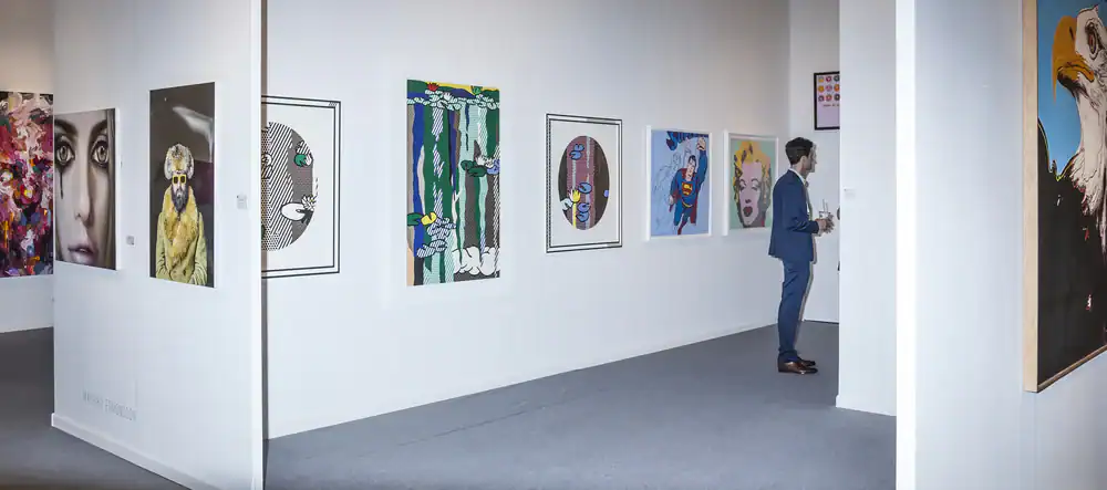 A man in a blue suit stands in a modern art gallery, observing colorful and diverse paintings hung on white walls. The artworks include abstract and figurative pieces, with one large eagle portrait visible on the right.