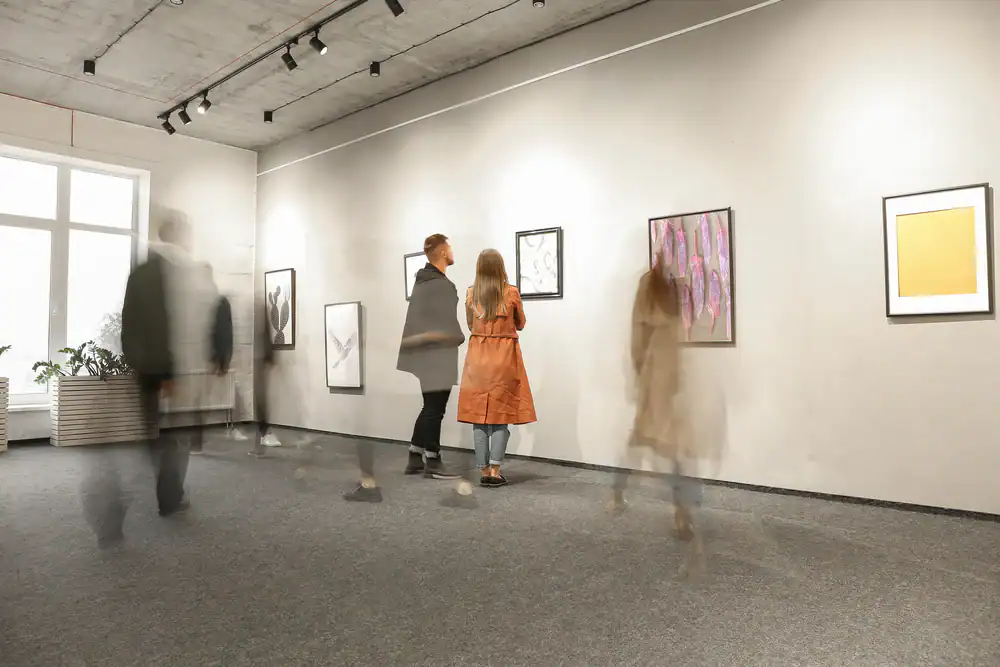 Several people view framed artwork on the walls of a modern art gallery. Some figures are blurred, showing motion, while two individuals stand still, closely observing a piece. The room has gray flooring and a concrete ceiling.