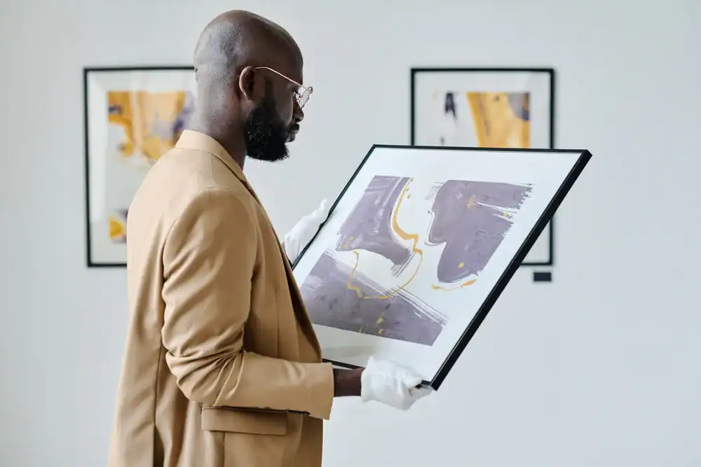 A man wearing a tan suit and white gloves examines a framed abstract painting with purple and yellow tones in an art gallery. Other abstract artworks are visible on the wall behind him.