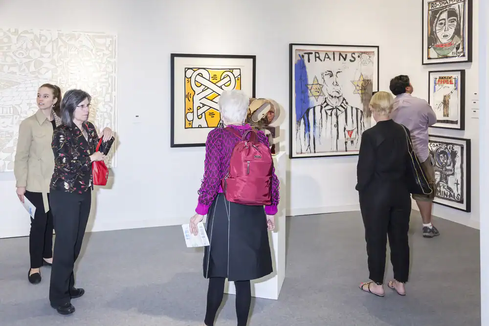 Several people view modern artworks displayed on white walls in a brightly lit gallery. Most are standing and observing the art, while one person leans in closer. The room appears calm and focused.