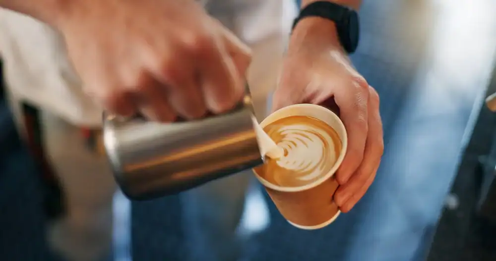 A person pours steamed milk from a metal pitcher to create latte art in a paper cup of coffee, holding the cup steady with their other hand at a bustling cafe NYC.