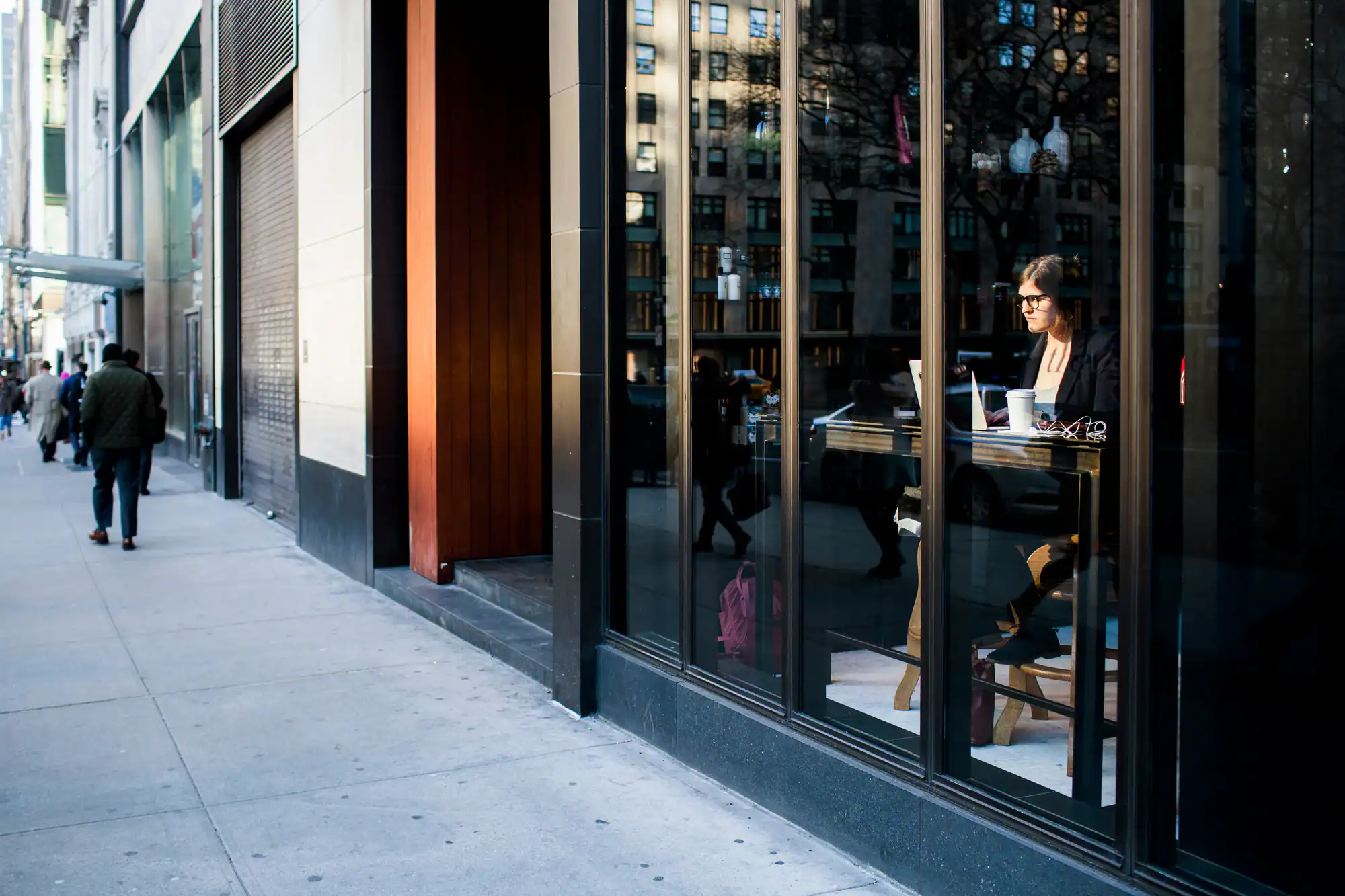A person sits alone at a tall table inside a cafe, working on a laptop by the window. The street outside is visible through large glass panes, with pedestrians walking by on the sidewalk.