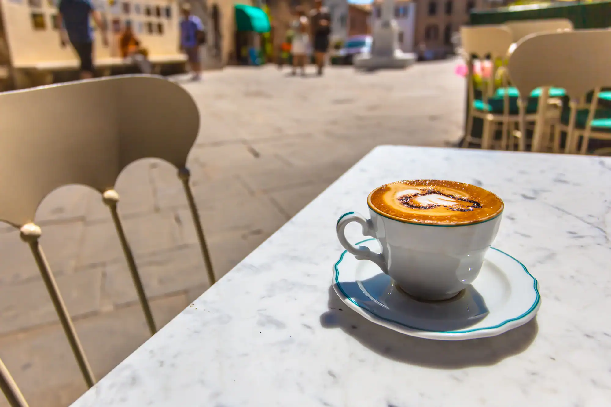 A cup of cappuccino with latte art sits on a marble table at an outdoor café, with blurred chairs, people, and buildings in the sunny background.