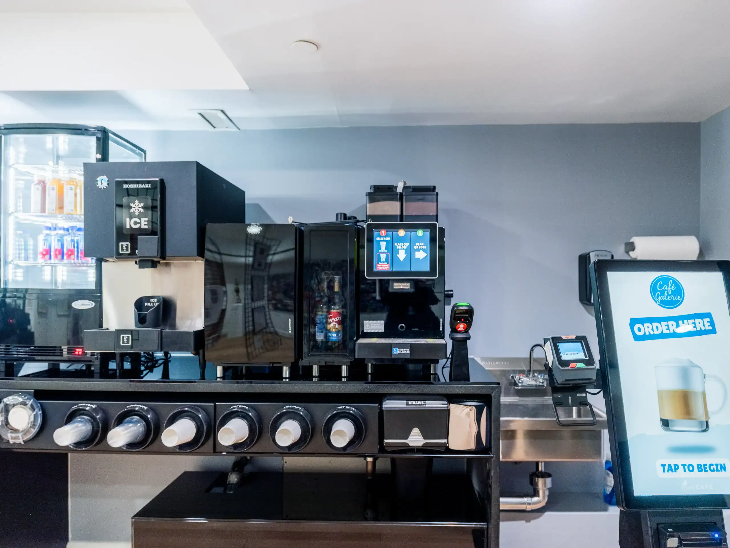 A self-serve drink station with an ice dispenser, coffee machines, drink dispensers, a refrigerated display with bottled drinks, and a touchscreen for ordering. The setup is clean and modern.