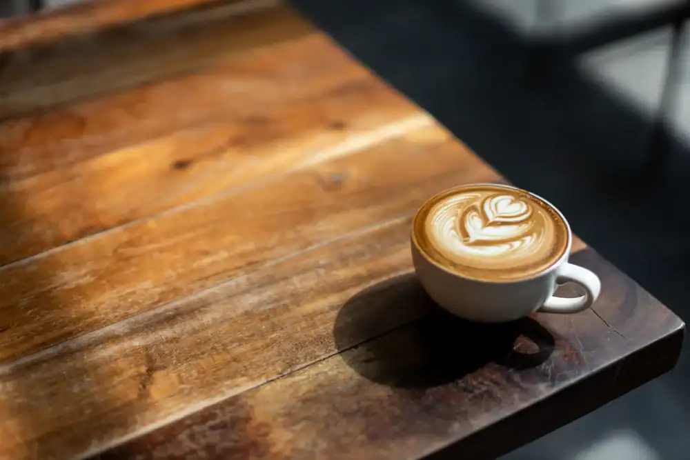 A cup of coffee with leaf-shaped latte art sits on a sunlit wooden table in a cozy cafe NYC, with light and shadows creating a warm, inviting atmosphere.