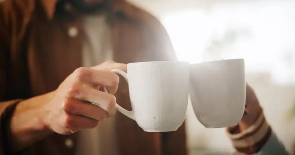 Two people clinking white ceramic mugs together in a toast, with a soft, warm light in the background. Only their hands and part of their torsos are visible.