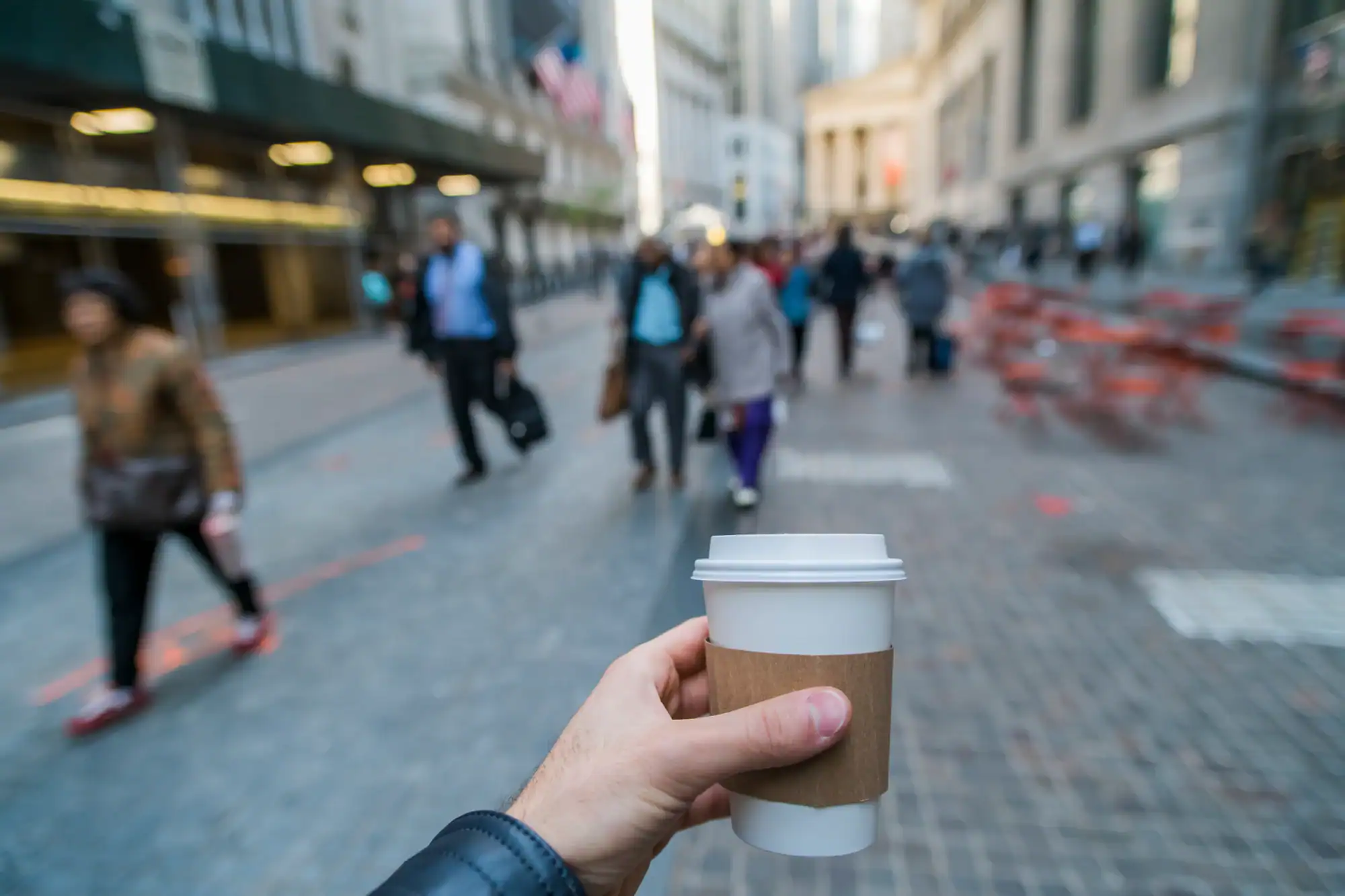 A hand holding a takeaway coffee cup in the foreground, with a busy city street and blurred pedestrians in the background. The scene appears to be in a financial district with tall buildings.