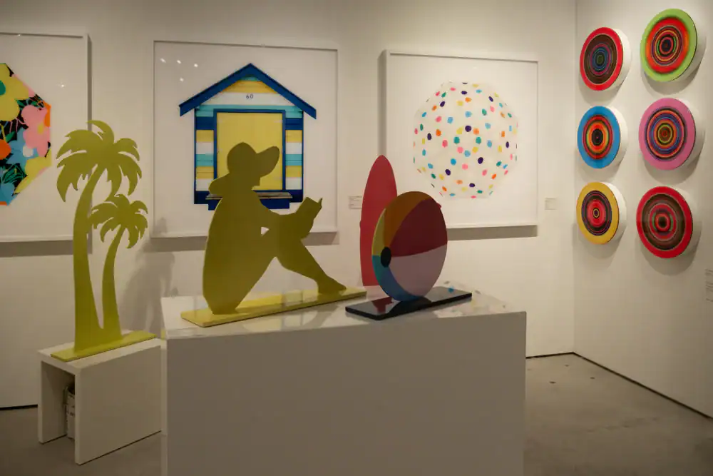 Colorful contemporary art exhibit featuring silhouettes of palm trees and a seated figure, a beach ball sculpture, a striped hut, and vibrant geometric paintings with circles and polka dots on the surrounding walls.