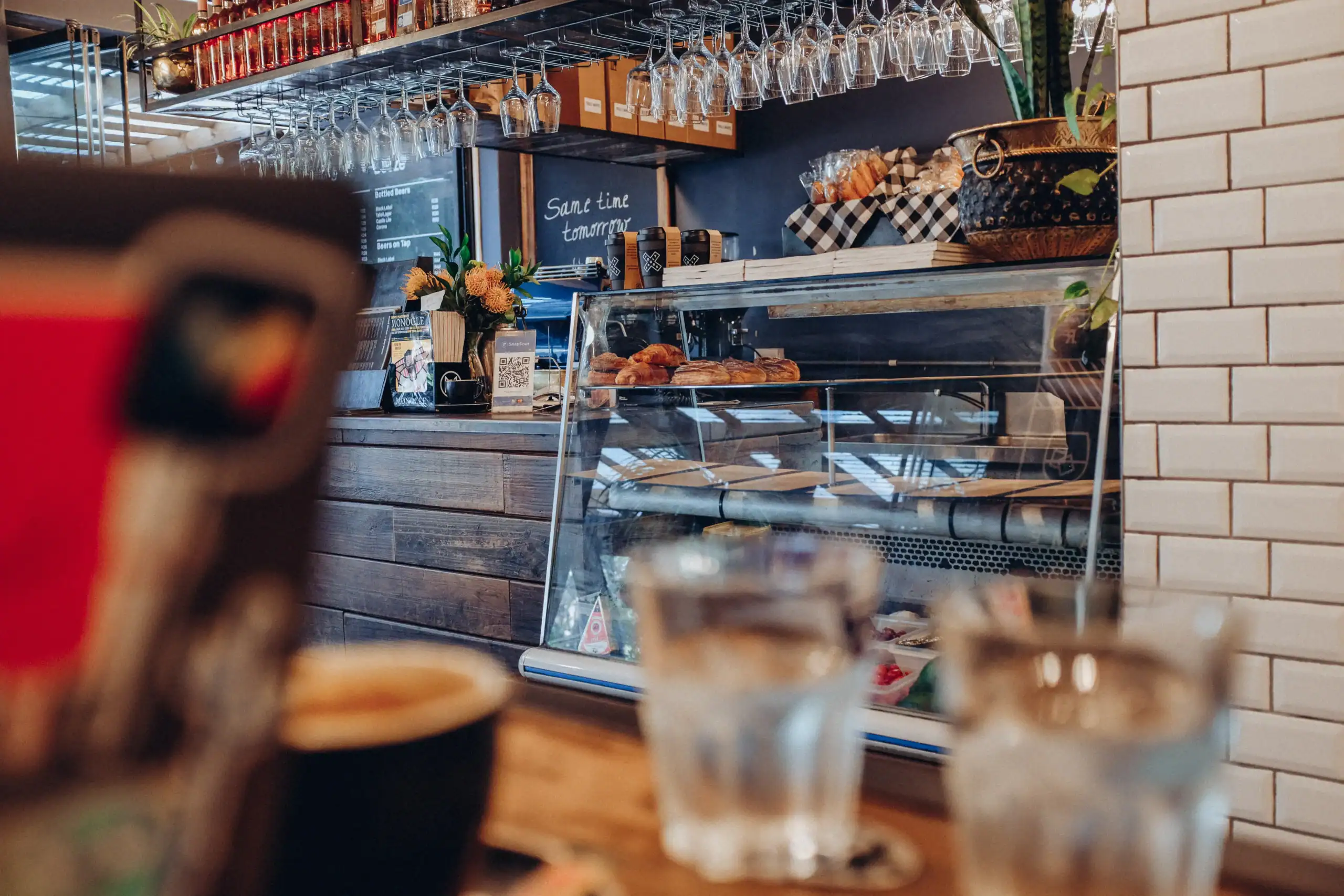 A cozy cafe interior with a glass display case filled with pastries, a wooden counter, hanging glasses, and shelves with various items. Two glasses of water and a coffee cup sit on a table in the foreground.