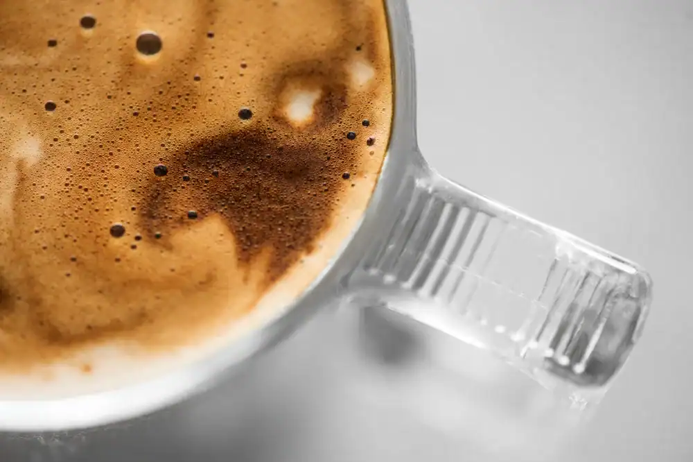 Close-up view of a glass mug filled with frothy coffee, showing bubbles and creamy foam on the surface, with the mug handle clearly visible on the right side.