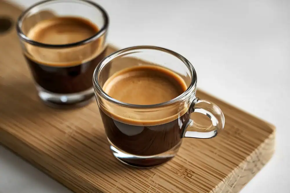 Two clear glass cups of espresso with crema on top sit on a light wooden board, with one cup in the foreground and the other slightly blurred in the background.