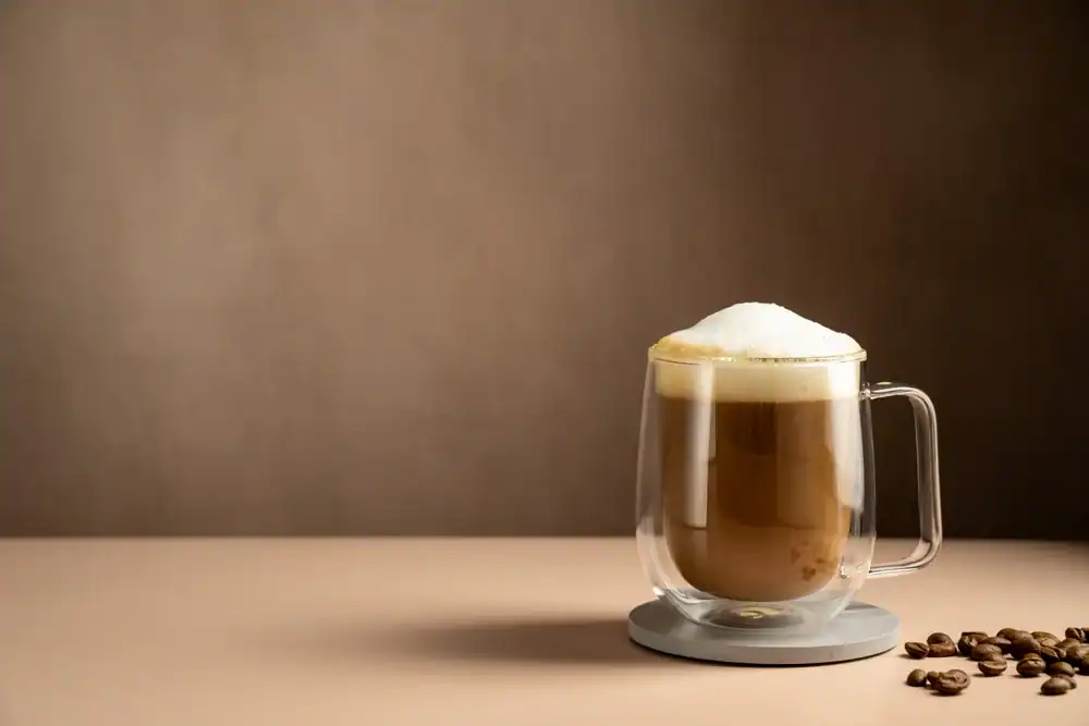 A glass mug of frothy cappuccino sits on a coaster, with a few coffee beans scattered nearby on a light-colored surface, evoking the cozy charm of a cafe NYC against a plain brown background.