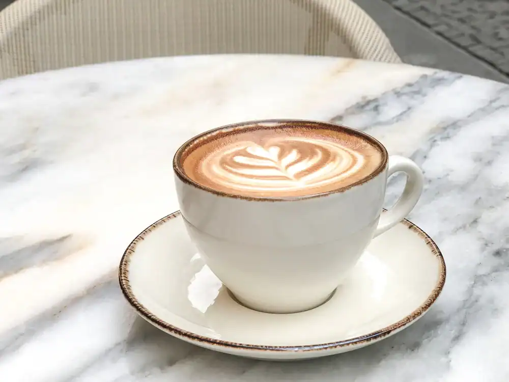 A white ceramic cup of cappuccino with leaf-shaped latte art sits on a matching saucer atop a round, white marble table—perfect for enjoying at a cozy cafe NYC style. A beige chair is partially visible in the background.