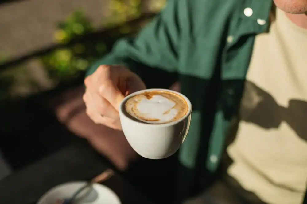A person in a green shirt and beige top holds a cup of coffee with latte art, seen from above in natural sunlight at a cozy cafe NYC. The white coffee cup rests on a saucer with a spoon on the table below.