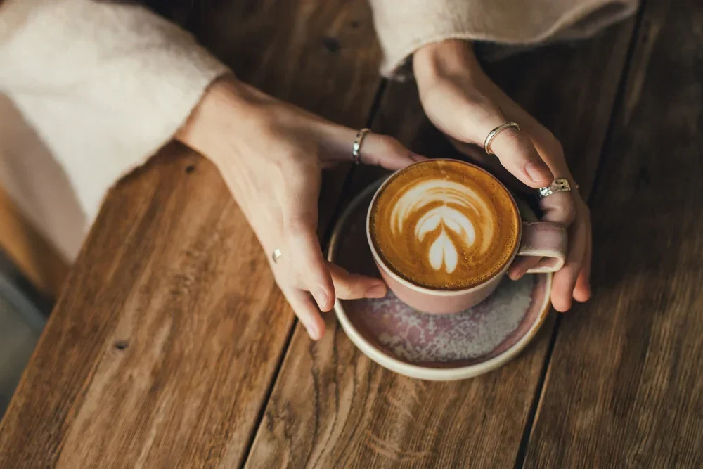 A person with several rings holds a cup of latte with leaf-shaped latte art, placed on a saucer on a rustic wooden table in a cozy cafe NYC setting.