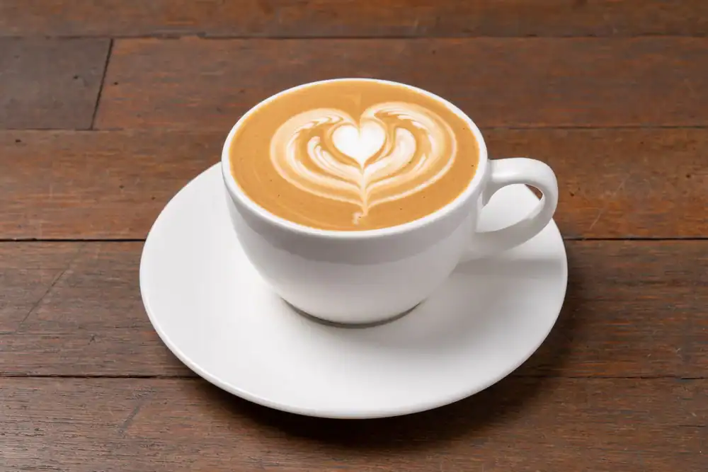 A white cup of coffee with heart-shaped latte art sits on a matching saucer atop a wooden table, capturing the cozy vibe of a cafe NYC morning.