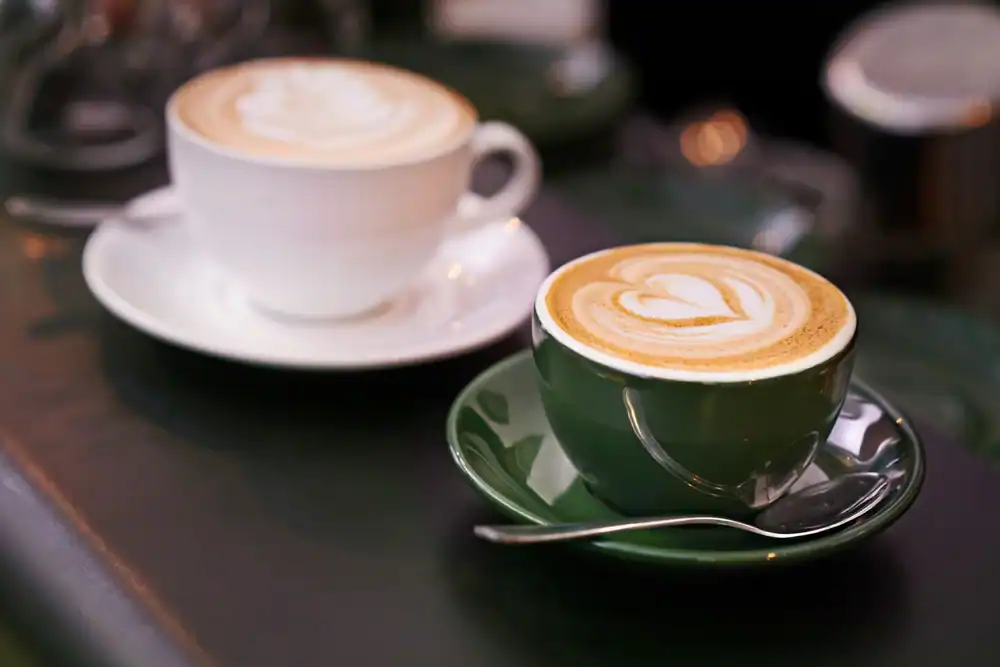 Two cups of cappuccino with heart-shaped latte art sit on saucers, one cup white and one cup green, each with a spoon beside them on a dark surface.