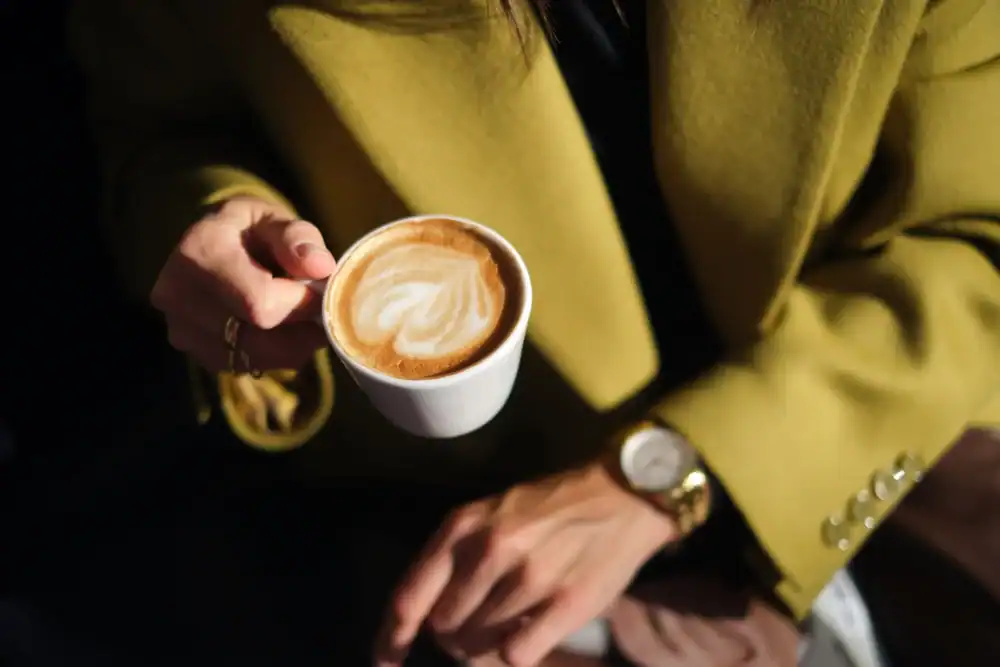 A person wearing a yellow coat holds a cup of cappuccino with latte art in one hand, and their other hand, adorned with a watch and rings, rests on their lap. The image is taken from above in soft lighting.