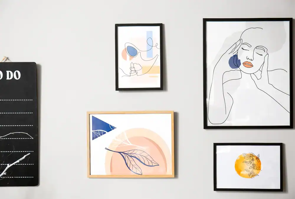 A gallery wall with four framed art prints, including abstract shapes, a minimalist line drawing of a person, stylized leaves, and a circular floral design, displayed on a light-colored wall next to a black to-do list board.