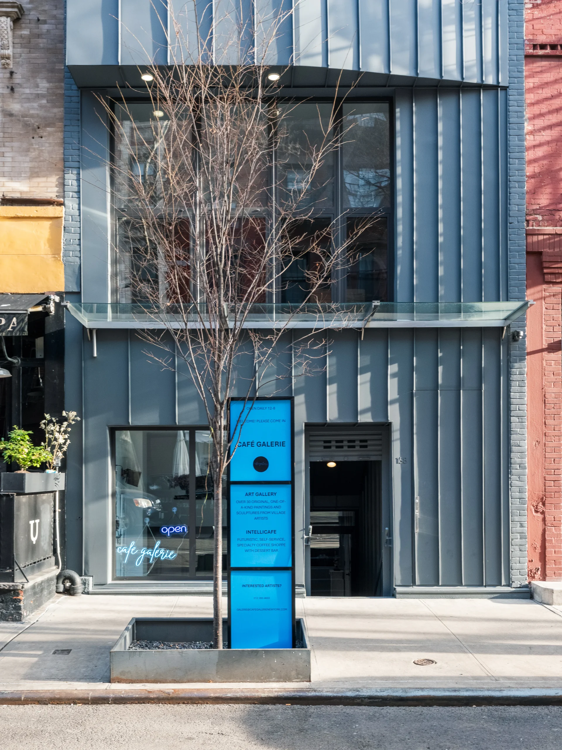A modern gray building with large glass windows and a blue vertical sign reading &ldquo;SAFE GALERIE&rdquo; stands on a city sidewalk, with a bare tree planted in front of the entrance.