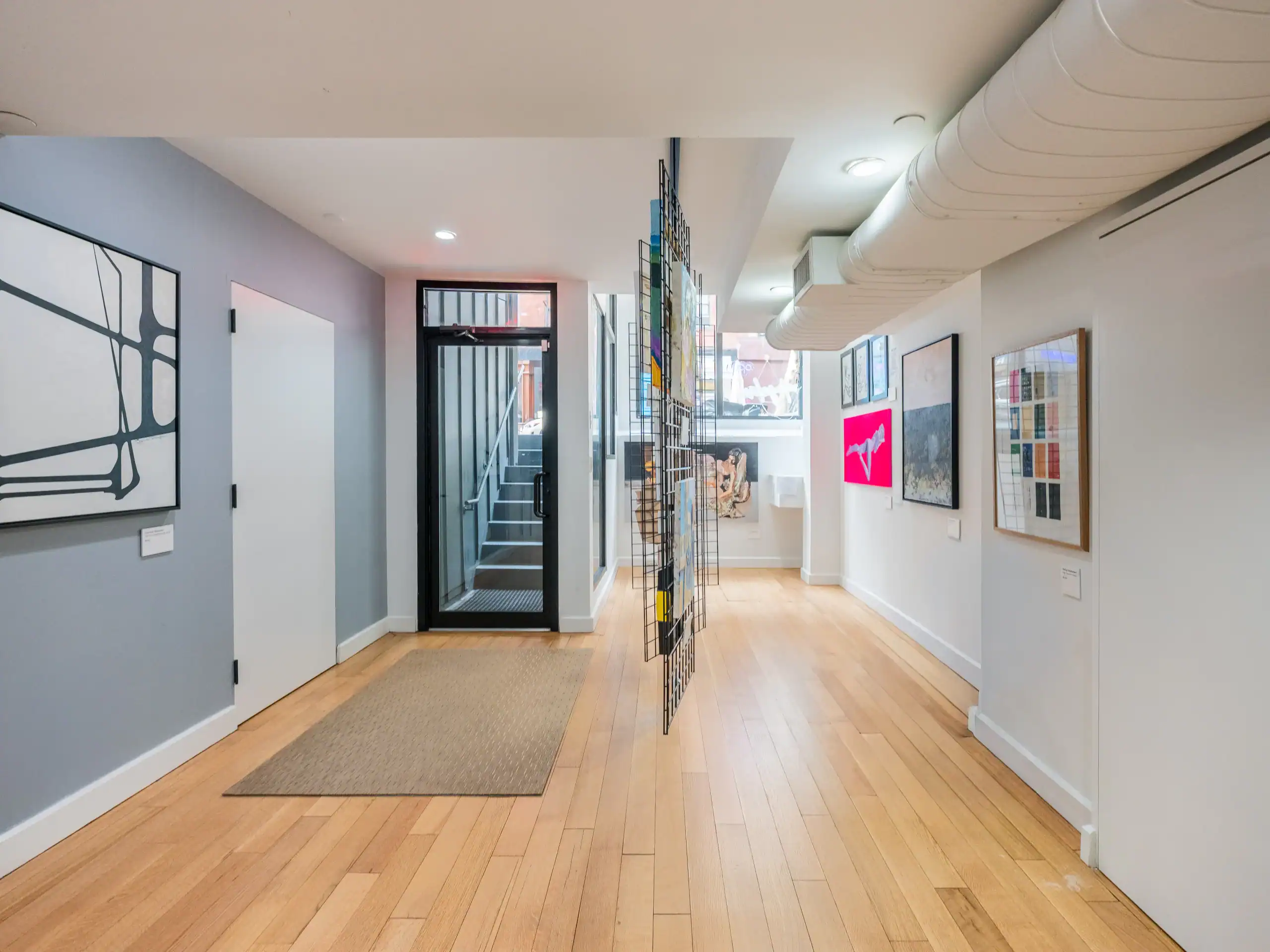 A modern hallway with light wood flooring, gray walls, abstract art, and a wall-mounted wire grid. A glass door at the end reveals stairs leading up, and a beige mat is placed near the entrance.