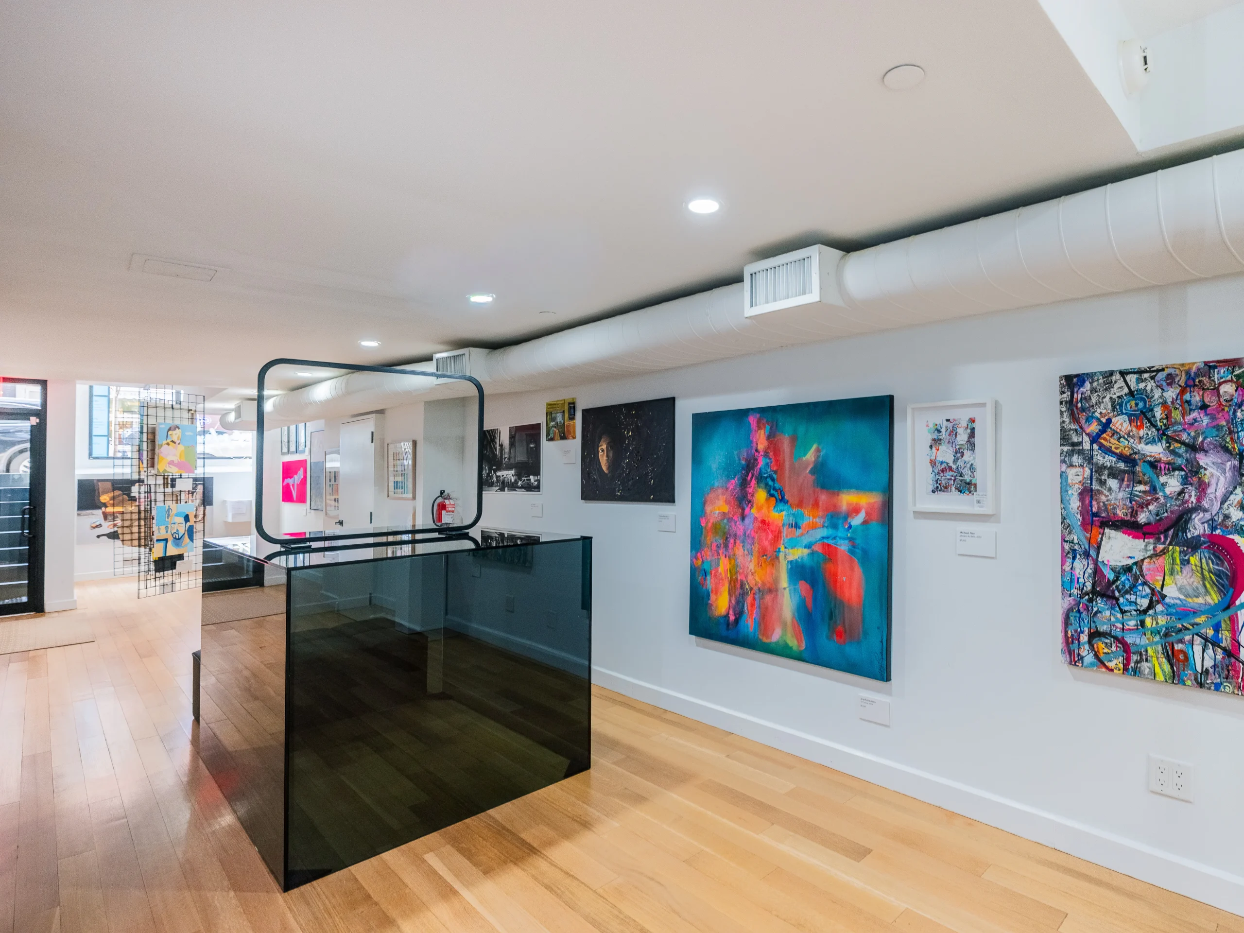 A modern art gallery with light wood floors, white walls, and colorful abstract paintings displayed. A glass and metal staircase is in the center, and bright lighting highlights the artwork.