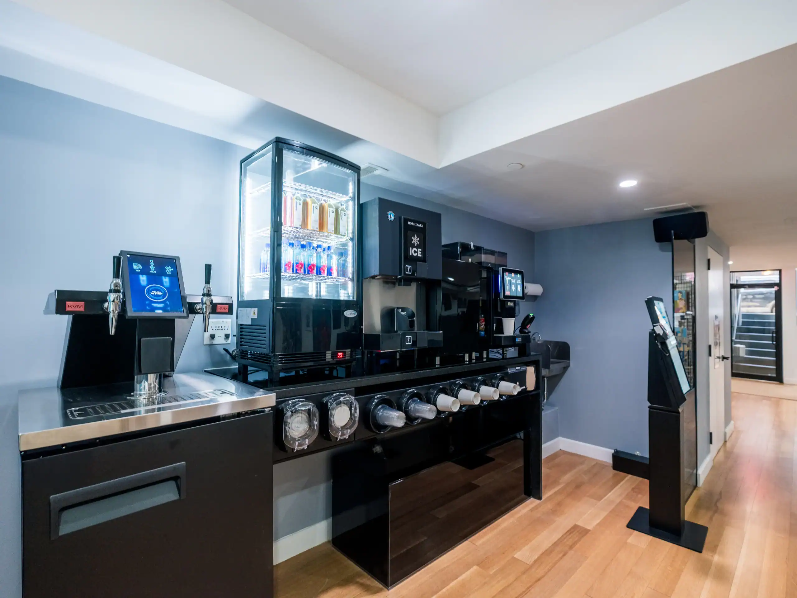 Self-service beverage station with a soda dispenser, ice machine, coffee maker, cup holders, touchscreen panel, and a refrigerated display case with drinks against a light blue wall on a wooden floor.