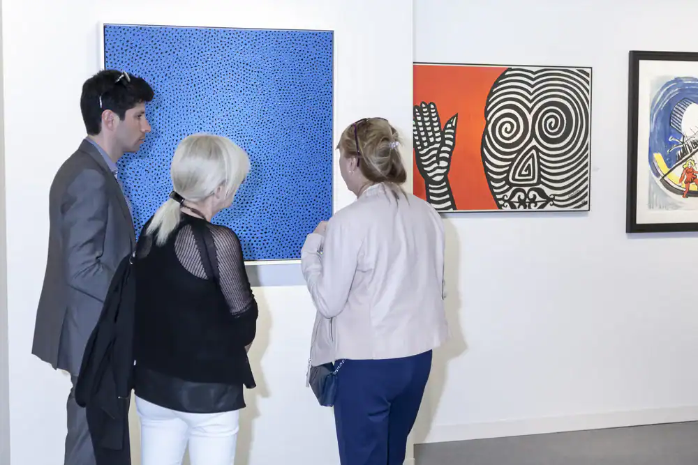 Three people stand in an art gallery, viewing colorful modern paintings on a white wall. One painting is blue with dots, another is red and black with a face, and a third features abstract shapes.