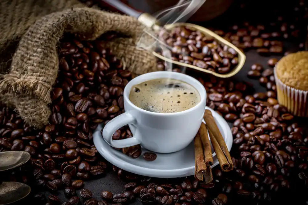 A steaming cup of black coffee in a white cup and saucer, with two cinnamon sticks beside it, surrounded by scattered coffee beans, a burlap sack, a muffin, and a coffee scoop&mdash;capturing the cozy vibe of a cafe NYC morning.