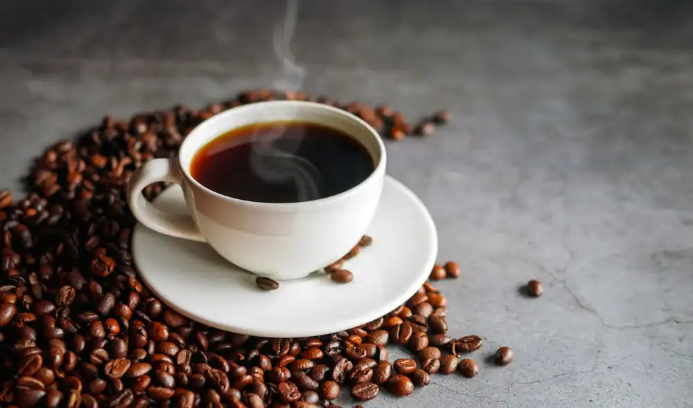 A steaming cup of black coffee sits on a white saucer, surrounded by scattered roasted coffee beans on a gray surface.