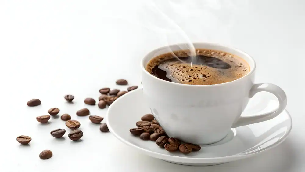 A steaming cup of black coffee in a white cup and saucer sits on a white surface, with scattered roasted coffee beans next to it.