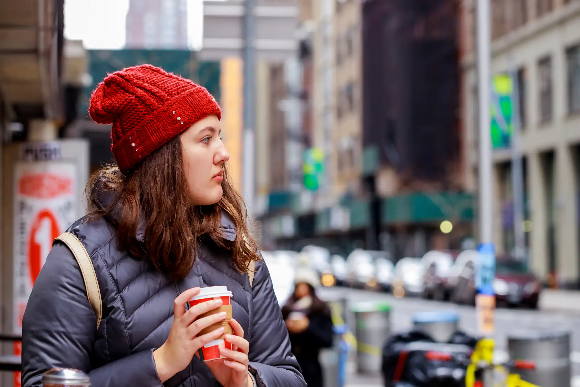 A woman in a red knit hat and gray winter coat holds a coffee cup and looks to the side while standing on a busy city street with blurred cars and buildings in the background.