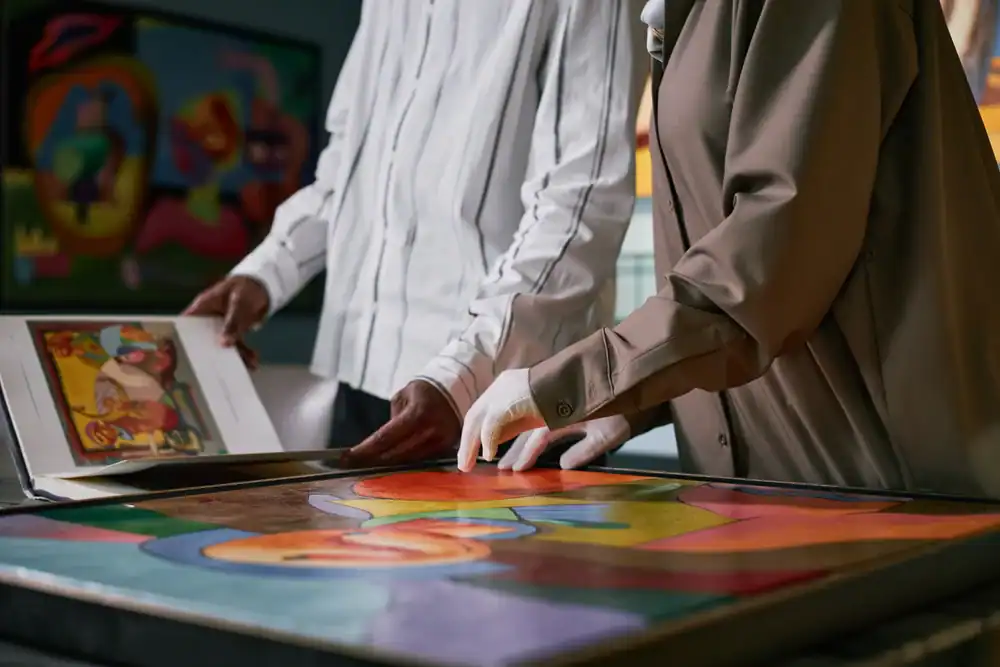 Two people examine colorful abstract artwork; one holds a book with art images, while the other, wearing white gloves, gently touches a framed painting on a table. Both are dressed in long-sleeved clothing.