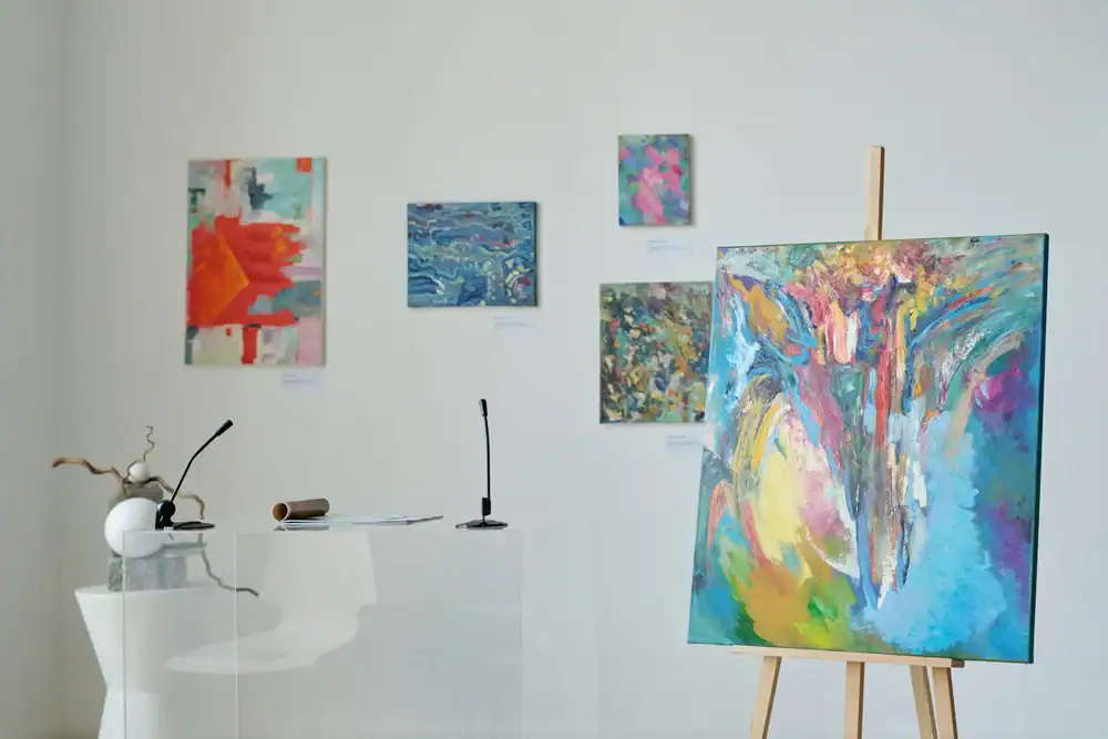 A modern art gallery features abstract paintings on white walls, with a large colorful canvas on an easel in the foreground, a clear podium with two microphones, and a white sculpture on a table nearby.