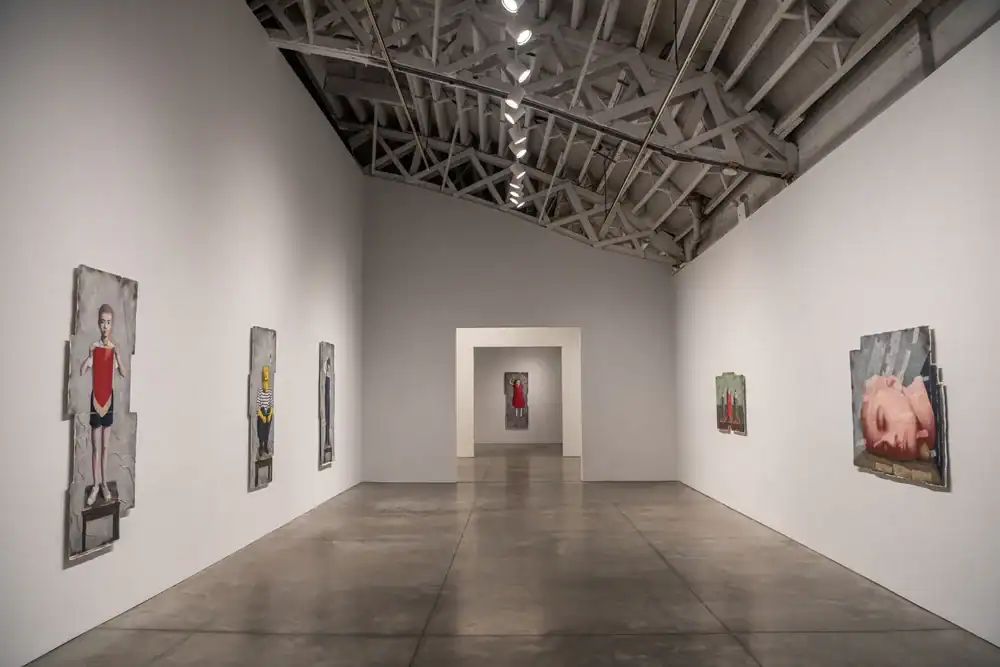 A modern art gallery with polished concrete floors and exposed beams displays figurative paintings on white walls, featuring people and objects. A doorway at the end reveals another painting in the adjacent room.