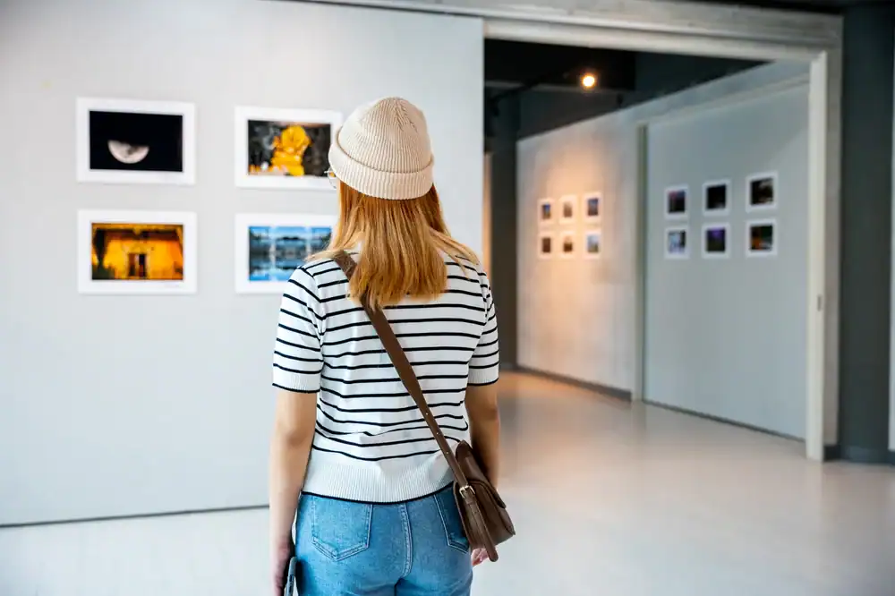 A woman with blonde hair, wearing a striped shirt, jeans, and a beige beanie, stands in a gallery viewing framed photographs displayed on white walls.