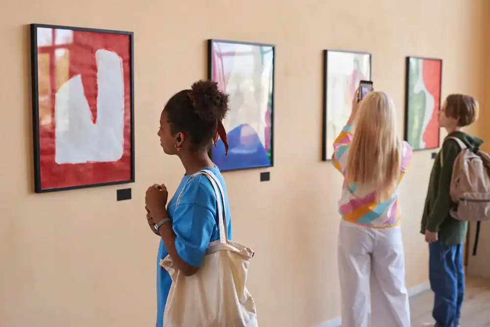 Three young people view colorful abstract art in a gallery. One person closely observes a painting, another takes a photo with a smartphone, and the third looks at the artwork on the wall.
