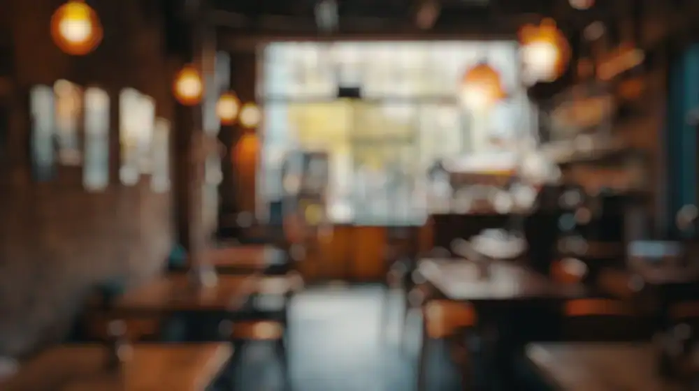 A blurred image of a cozy café interior with wooden tables, chairs, warm lighting, and a large window in the background.