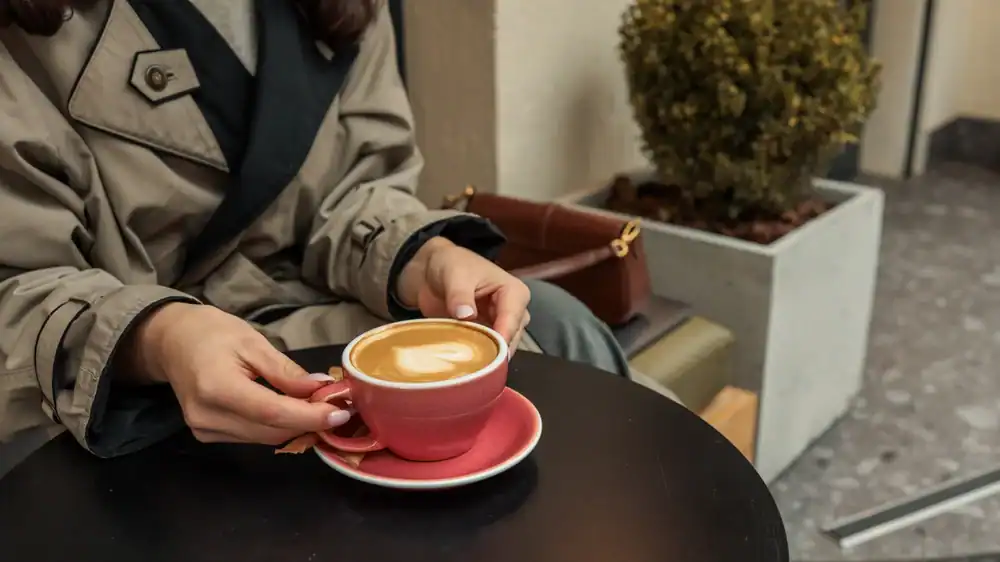 A person in a beige trench coat holds a pink cup of coffee with latte art, sitting at a black table outdoors. A purse and a potted plant are visible in the background.