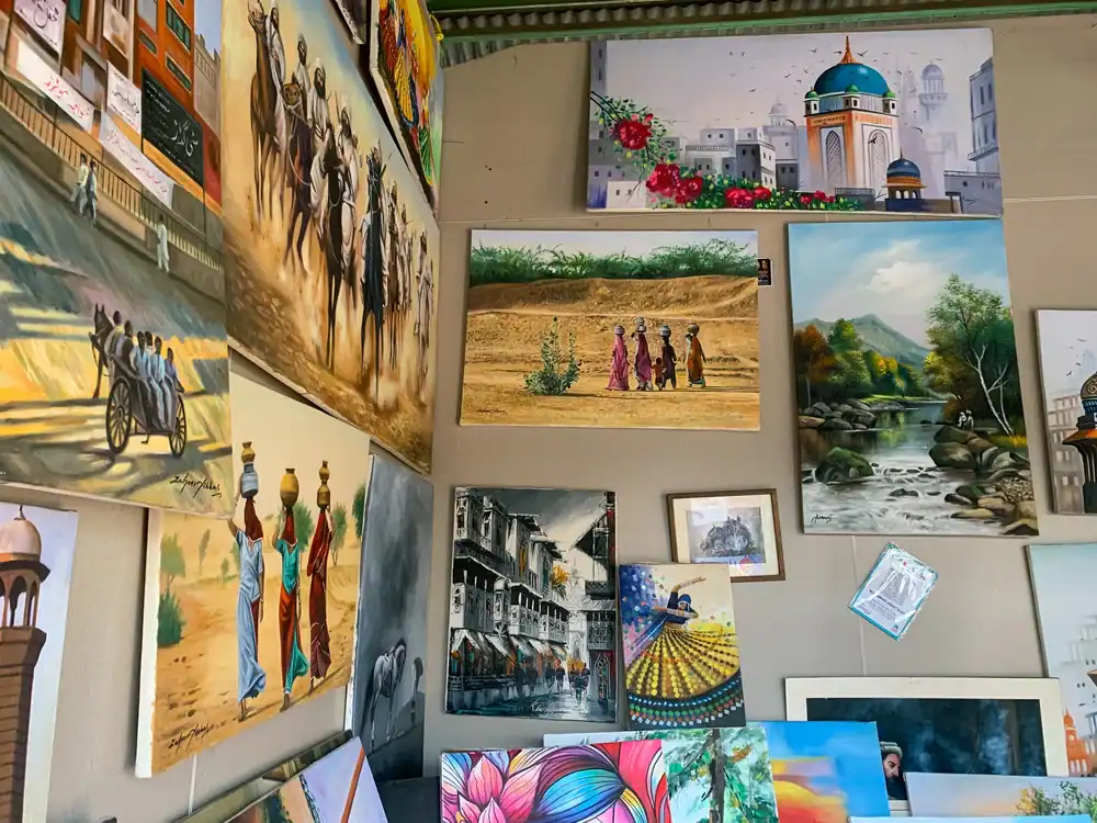 A wall displays colorful paintings, including scenes of people, city streets, nature, horse riders, and architectural landmarks, arranged closely together in a vibrant, artistic setting.