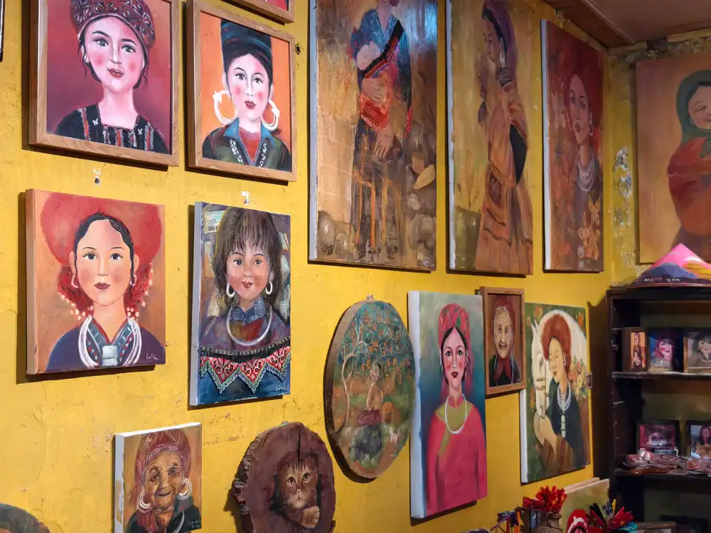 Colorful portraits of women in traditional attire are displayed on a yellow wall in an art gallery or shop, along with a few masks and small sculptures on a shelf beneath the paintings.