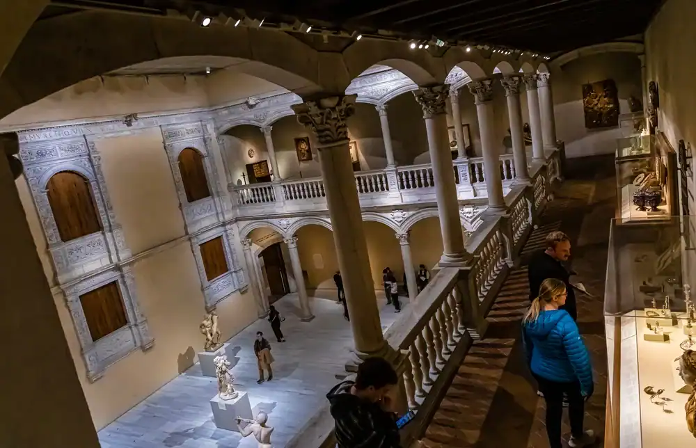 A view inside a museum with classical architecture, tall columns, and balconies. Several people are observing sculptures and displays, while soft lighting highlights the artwork and ornate details of the space.