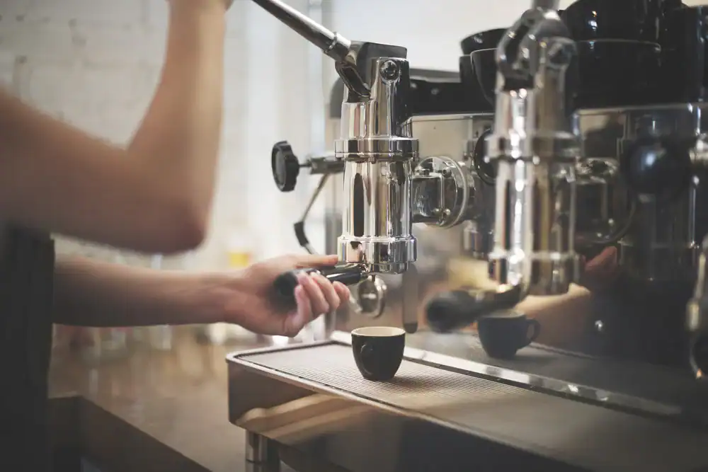 A person is using a professional espresso machine to make coffee, holding a portafilter and placing a small black cup under the spout as the espresso pours out.