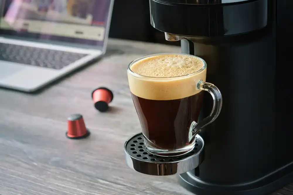 A glass mug filled with freshly brewed coffee sits on a coffee machine drip tray, with coffee pods and an open laptop in the blurred background on a wooden table.