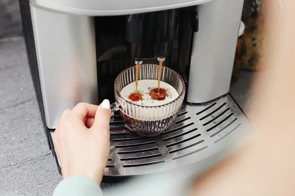A person holds a glass cup under a coffee machine as it brews coffee, with two streams of espresso pouring into the cup on a metal drip tray.