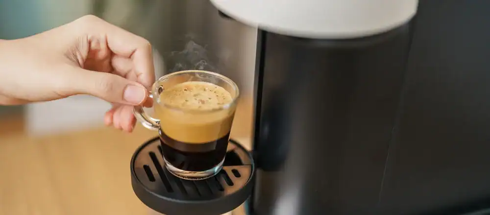 A hand holding a small glass cup of freshly brewed espresso with frothy crema, placed on the drip tray of a modern coffee machine. Steam is rising from the hot coffee.