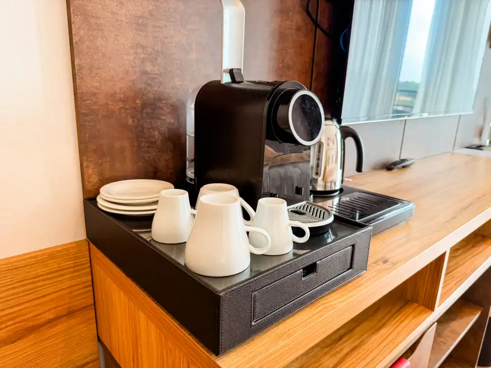 A coffee station on a wooden counter with a coffee machine, kettle, stacked white saucers, and four white cups arranged neatly on a black tray. A large mirror and windows are visible in the background.